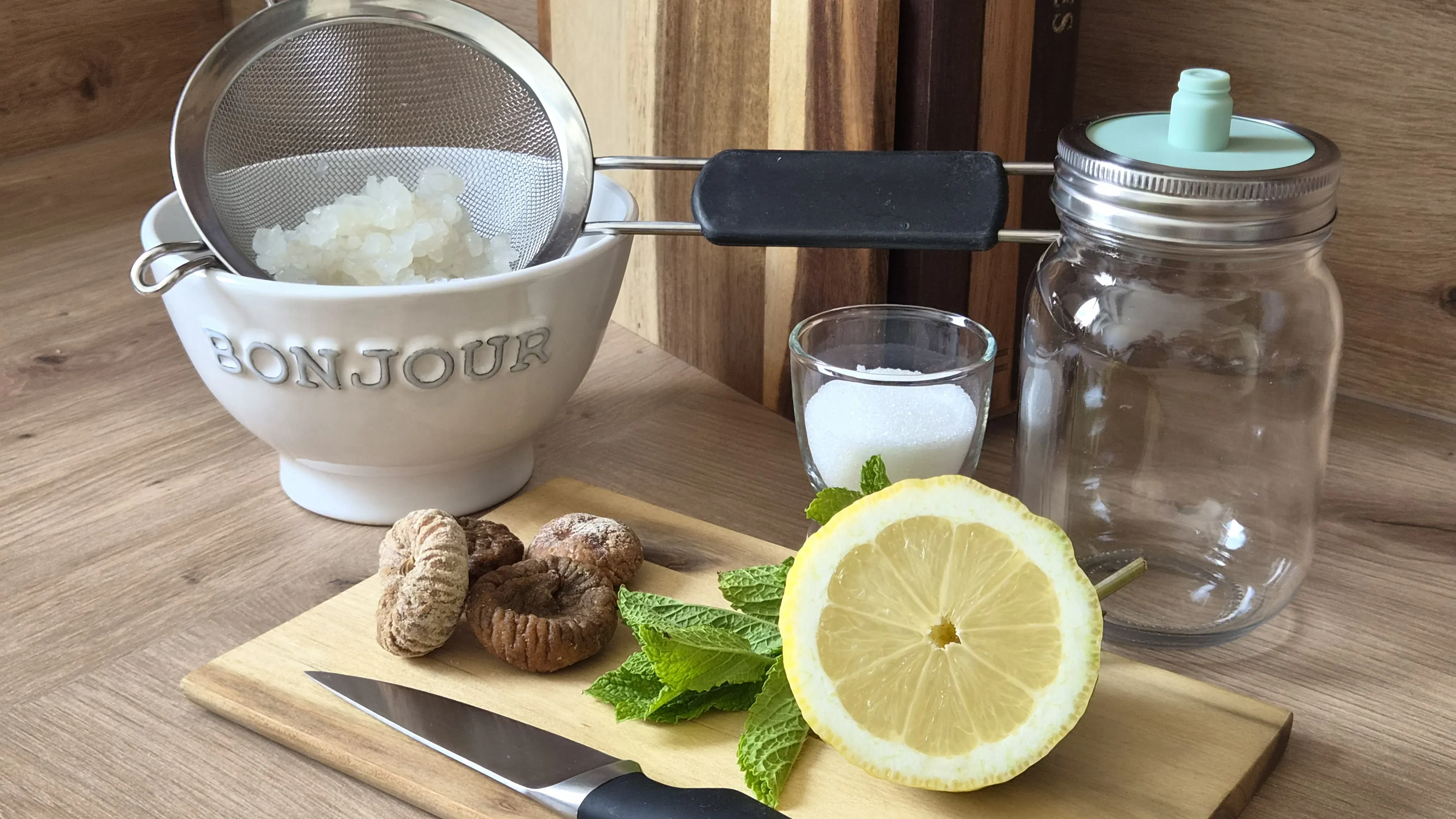 Zutaten und Zubehör für Wasserkefir: ein Sieb mit Wasserkefir-Kristallen über einer Schale, ein Glas Zucker, ein Bügelglas, getrocknete Feigen, Minze, eine halbe Zitrone und ein Messer auf einem Holzbrett